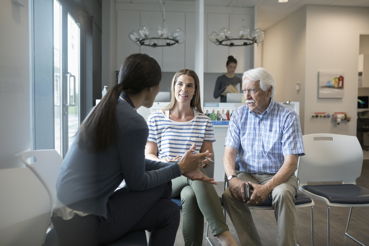 Female doctor talking with senior male patient and daughter in clinic waiting room